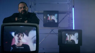 Ein Screenshot aus dem Musikvideo von Alphaville symphonischer Neuauflage ihres Hits "Big In Japan" zeigt Sänger Marian Gold auf einen Fernseher gestützt, auf dem das Originalvideo aus dem Jahr 1984 zu sehen ist.