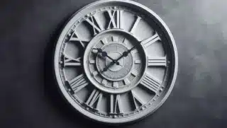 Das Bild zeigt eine Uhr mit Ziffernblatt und Uhrzeigern an einer dunkelgrauen Wand.