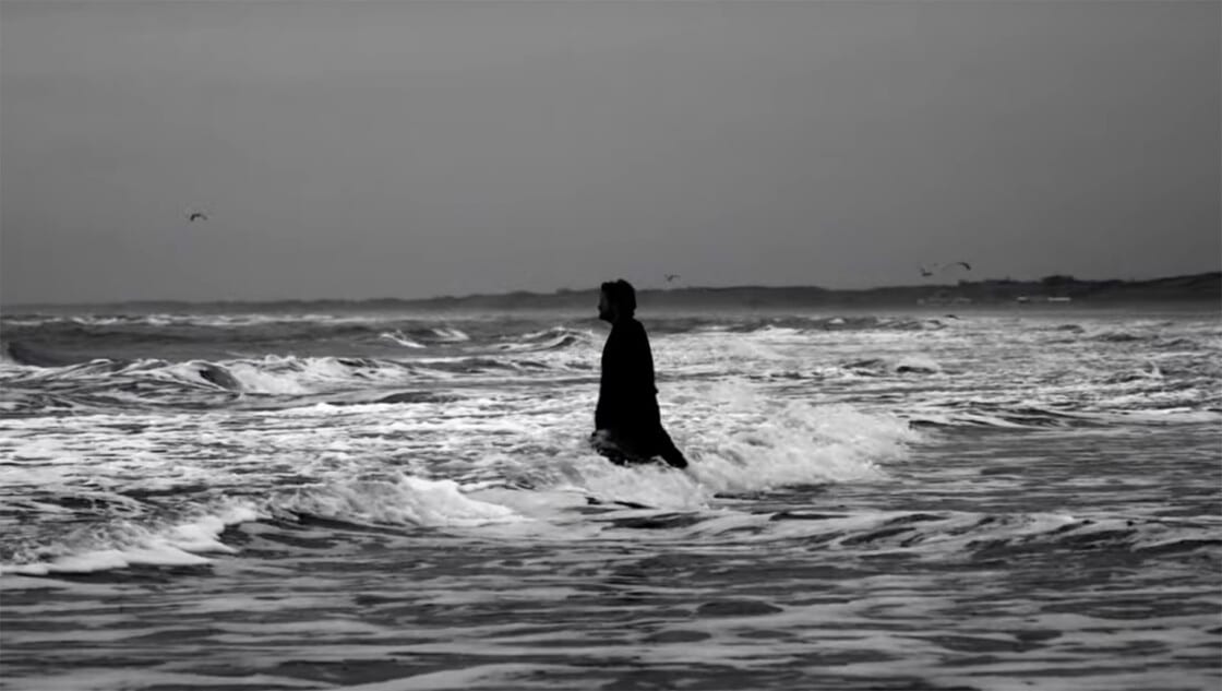 Der Videoausschnitt zeigt eine Schwarzweiß-Aufnahme mit einem Mann, der in die Schaumkronen eines aufgewühlten Meeres steigt.